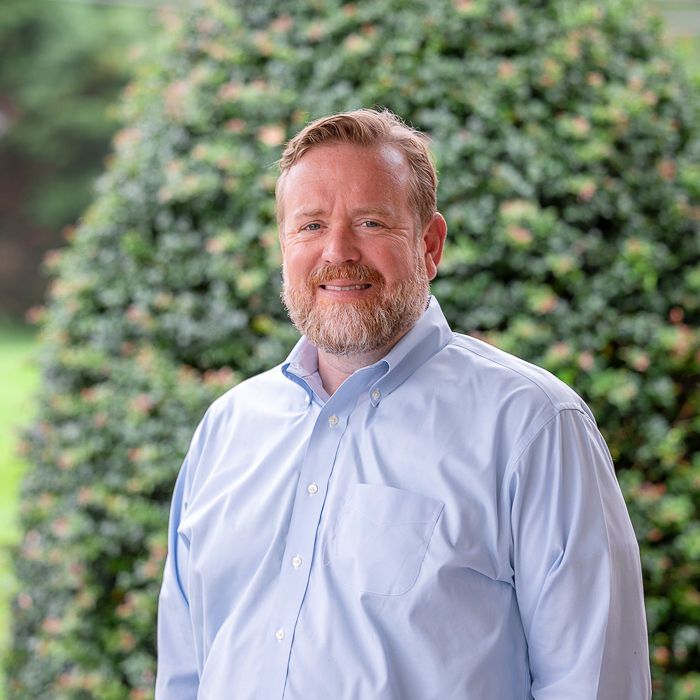 Smiling man in a light blue shirt standing outdoors before green foliage