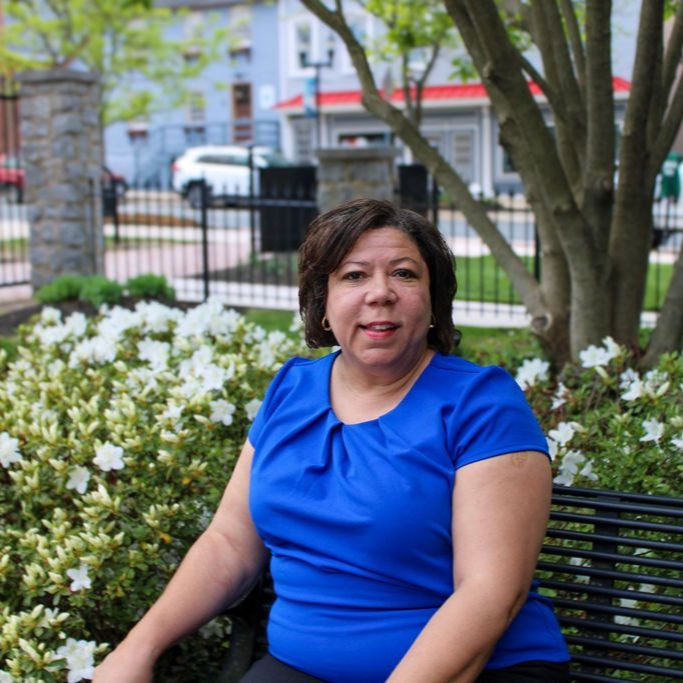 Woman in a blue patterned blouse and navy pants standing on a porch by a stone railing.