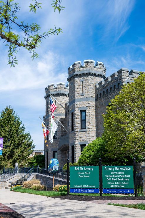 Stone castle entrance with flags and green signboards under a blue sky, framed by trees.