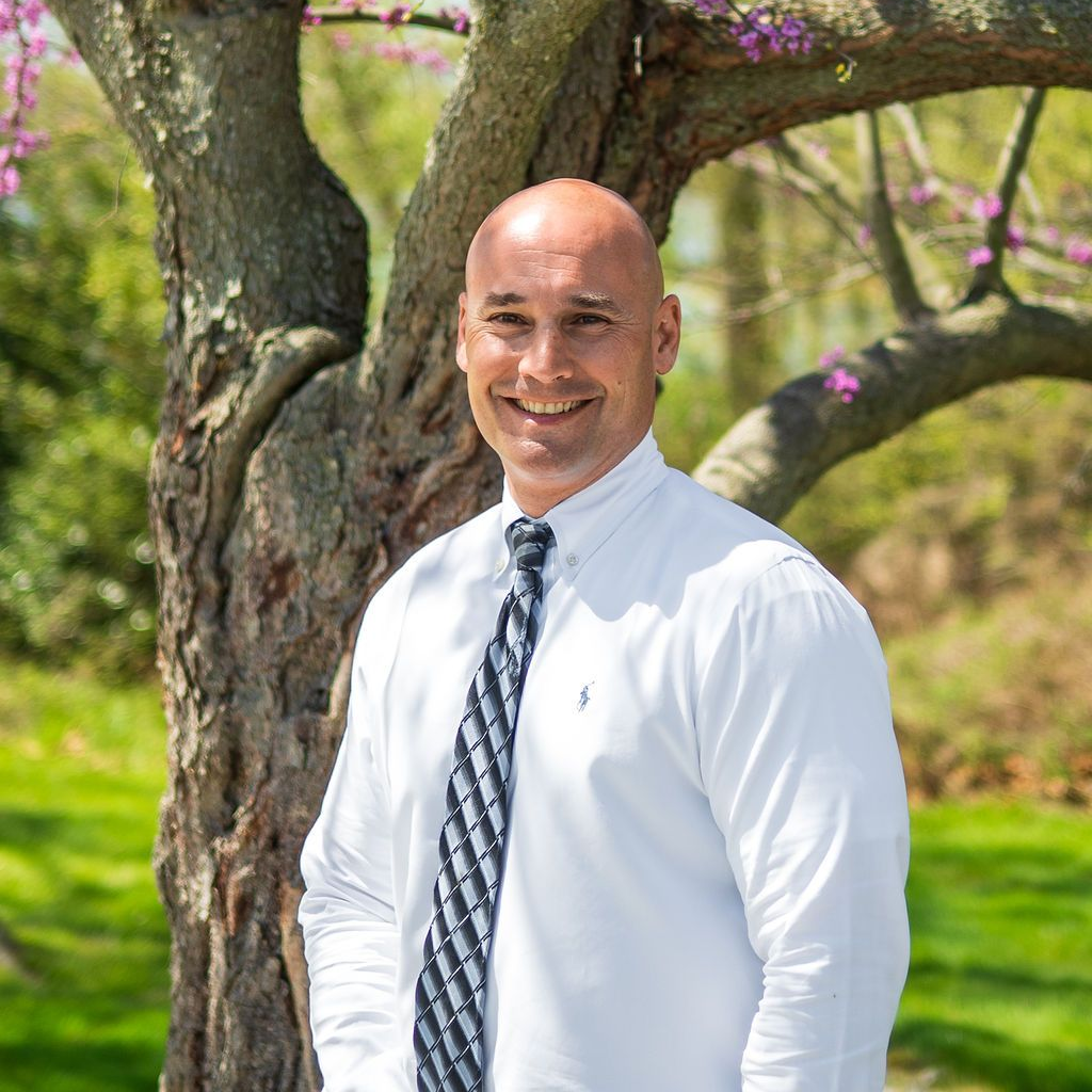 Smiling man in a white shirt and patterned tie standing by a blossoming tree outdoors
