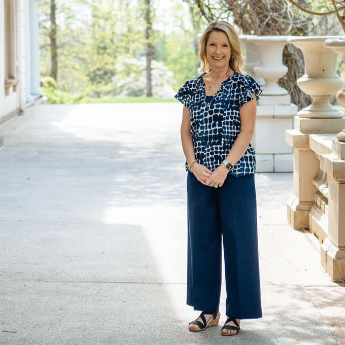 Woman in a blue patterned blouse and navy pants standing on a porch by a stone railing.