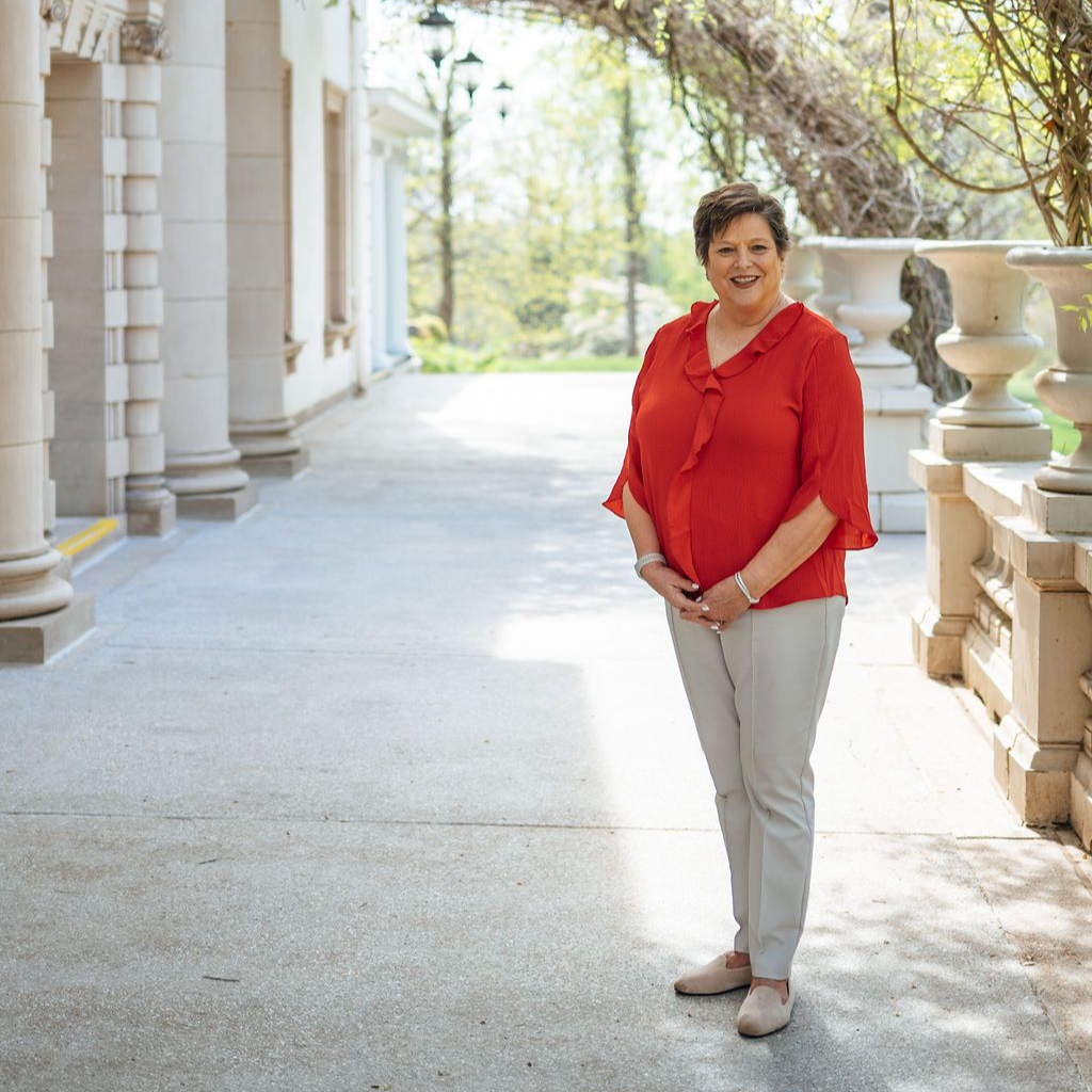 Person in a red blouse and beige pants standing on a sunlit colonnade walkway
