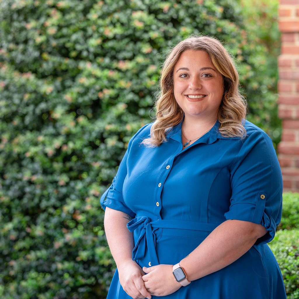 Smiling woman in a blue dress standing outdoors against a leafy green background
