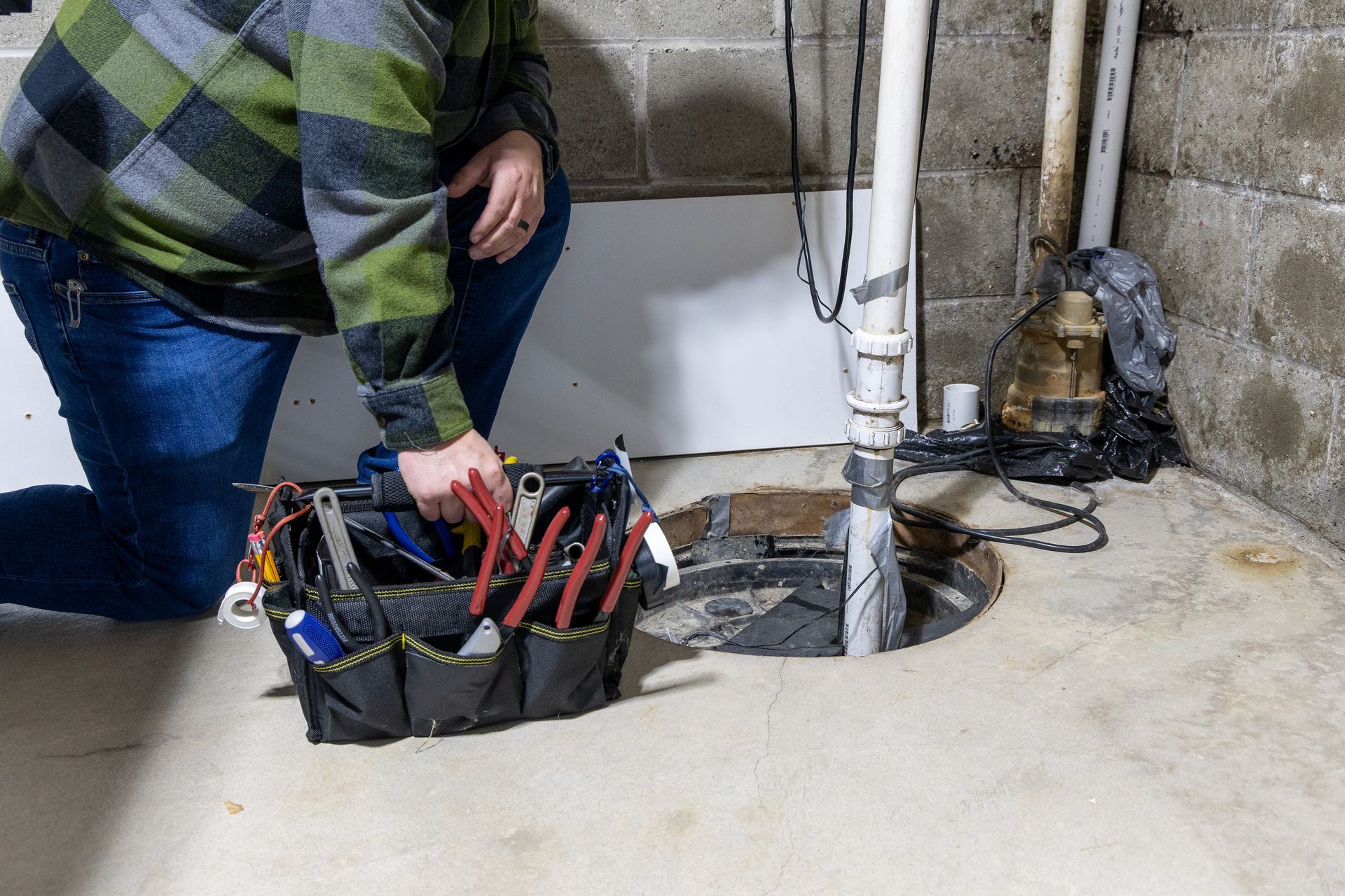 A technician with a tool bag kneeling beside a sump pump pit.