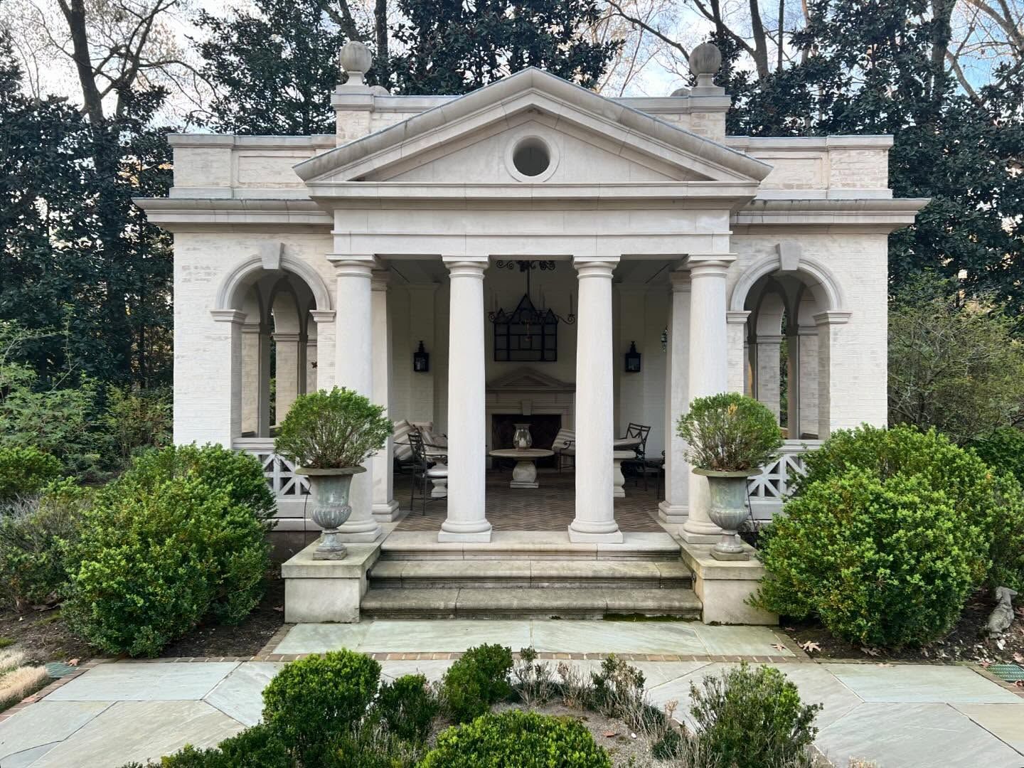 Stone pavilion with columns and steps, surrounded by shrubs, in a garden setting.