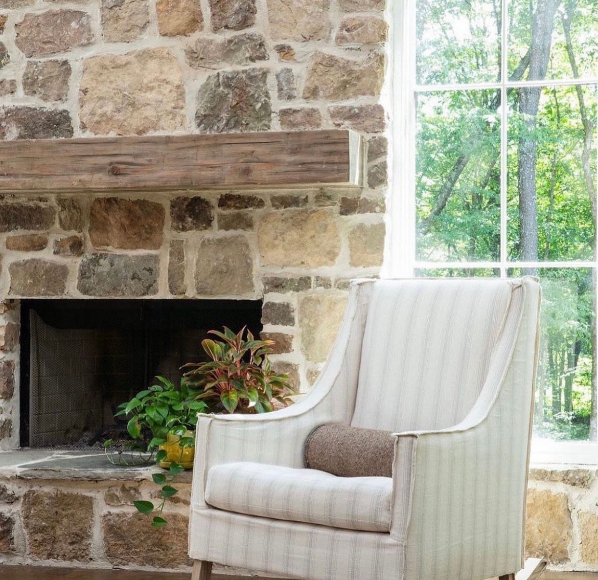 Stone fireplace with wooden mantel, next to a striped armchair and window overlooking a forest.