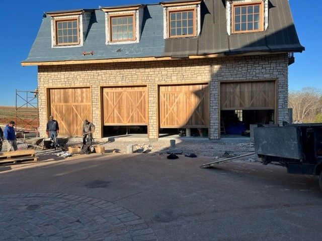 Three-bay garage with wooden doors, stone exterior, and dormers. Construction workers nearby.