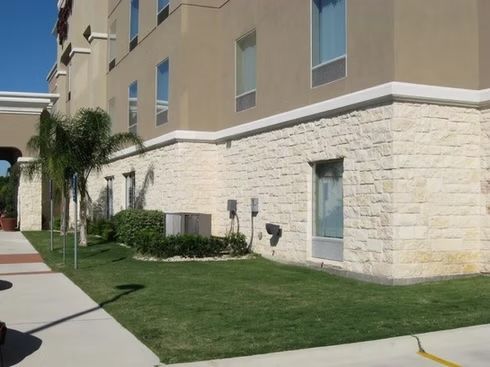 Exterior of a tan hotel building with stone base, green grass, and small trees on a sunny day.