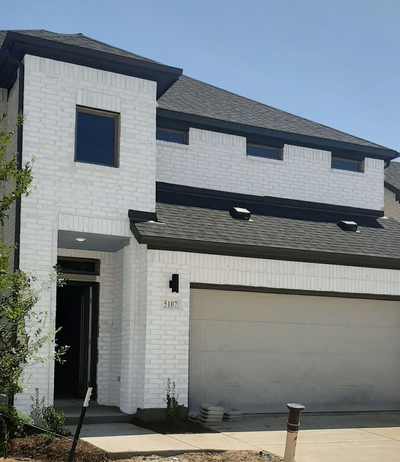 Two-story white brick house with black trim, dark roof, and gray garage door on a sunny day.