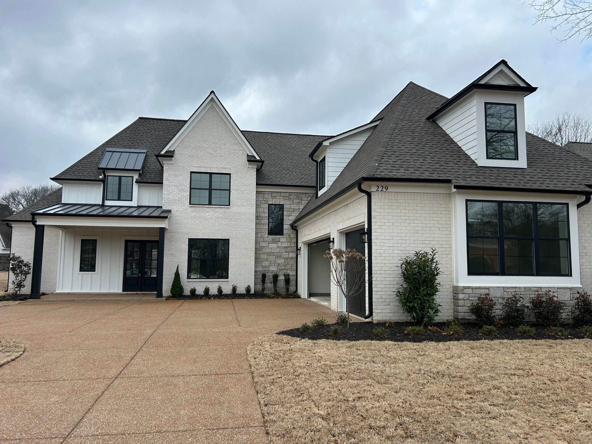 Two-story white brick house with dark windows, a brown roof, and a gravel driveway under an overcast sky.
