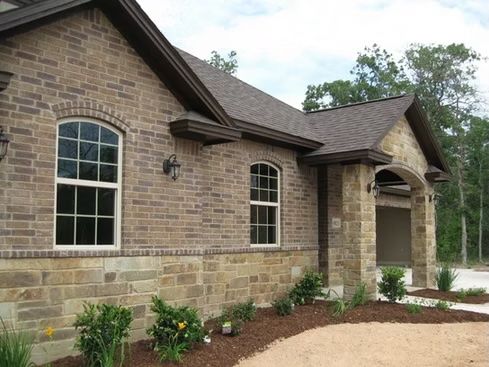 Brick house with arched windows and brown roof, set in a landscape with shrubs and gravel driveway.