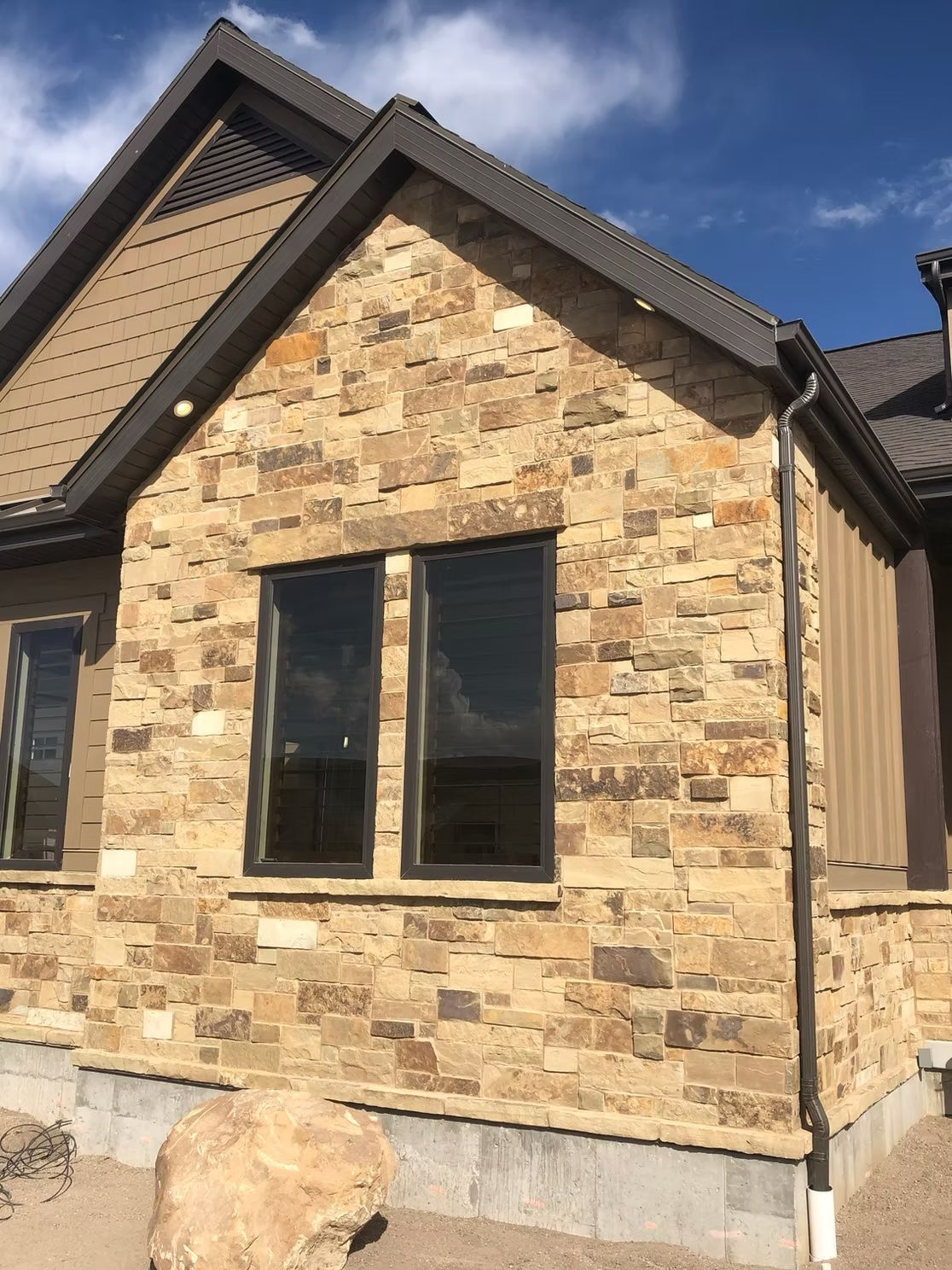 Tan stone facade of a house with dark framed windows and a brown roof against a blue sky.
