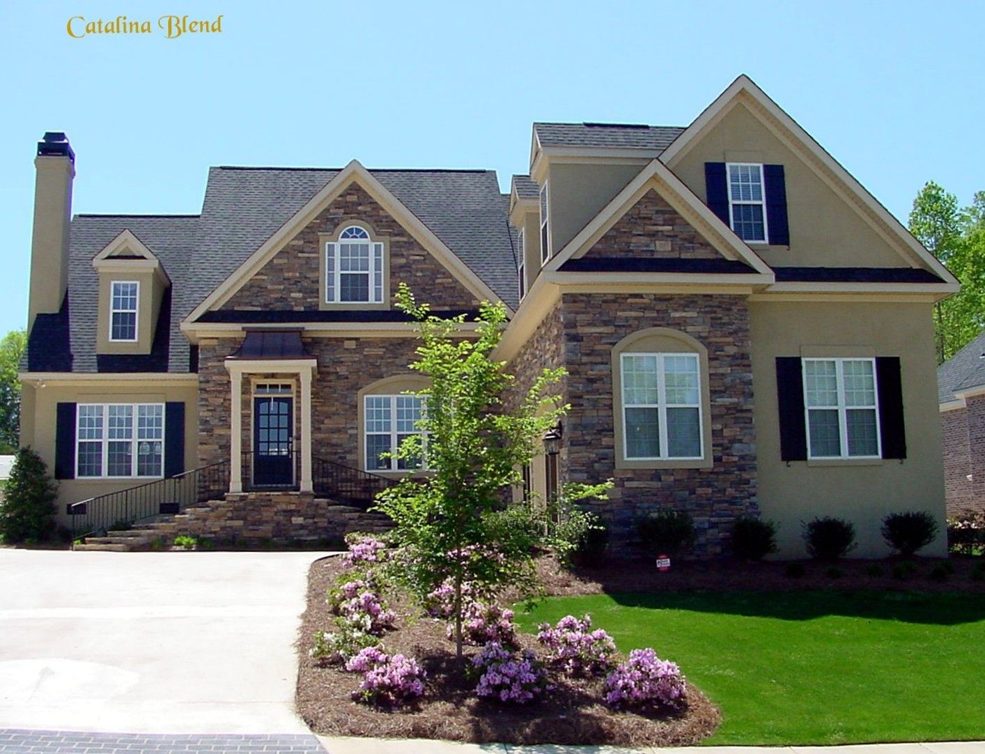 Multi-story house with stone and tan facade, black shutters, green lawn, and blue sky.