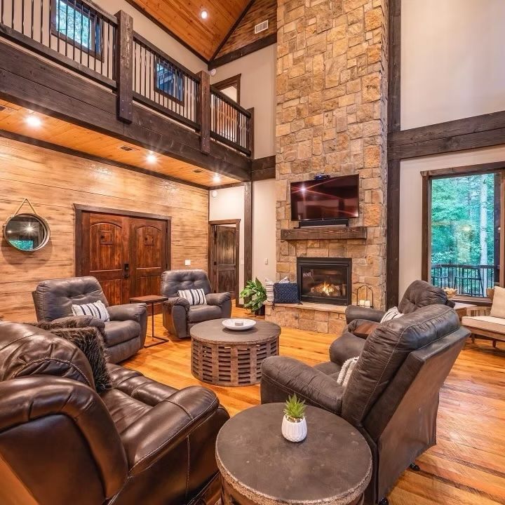 Living room with stone fireplace, wooden beams, leather chairs, and a second-floor balcony.