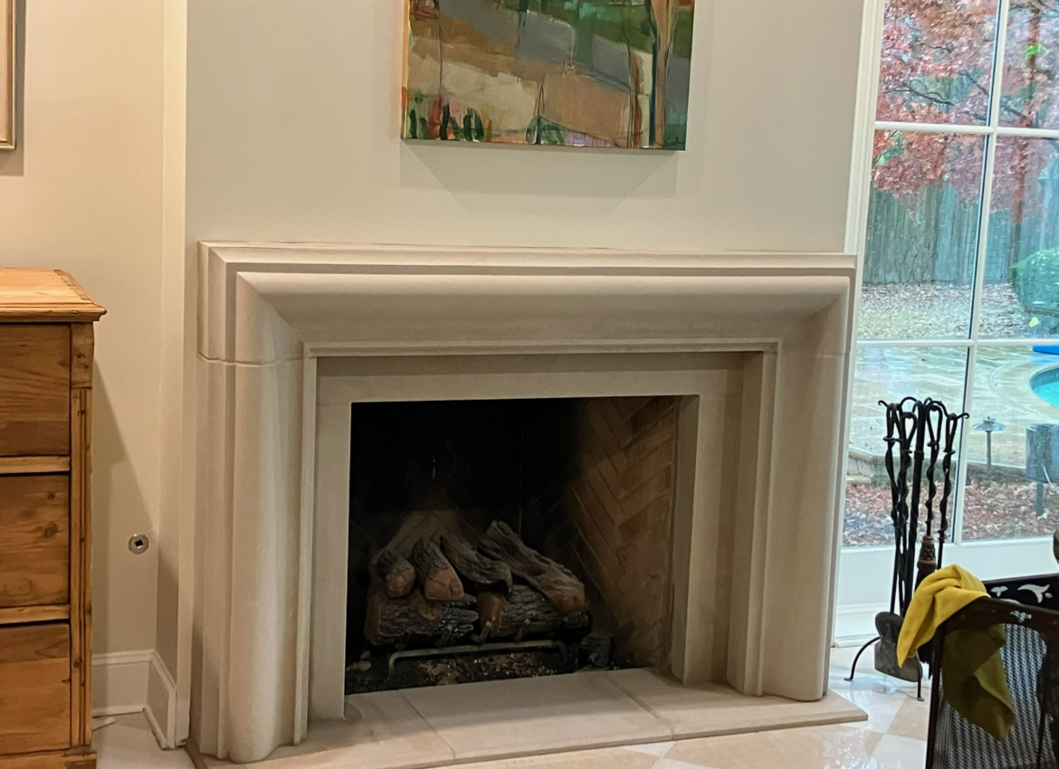 Fireplace with white mantel, artwork above, logs inside. Window view, trees outside.