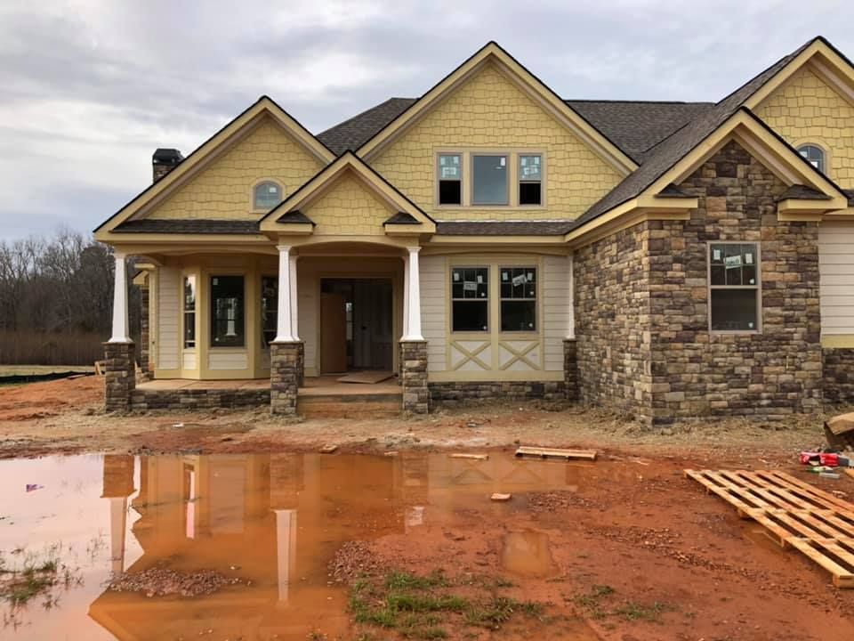 A house under construction with stone and yellow siding, surrounded by red mud.
