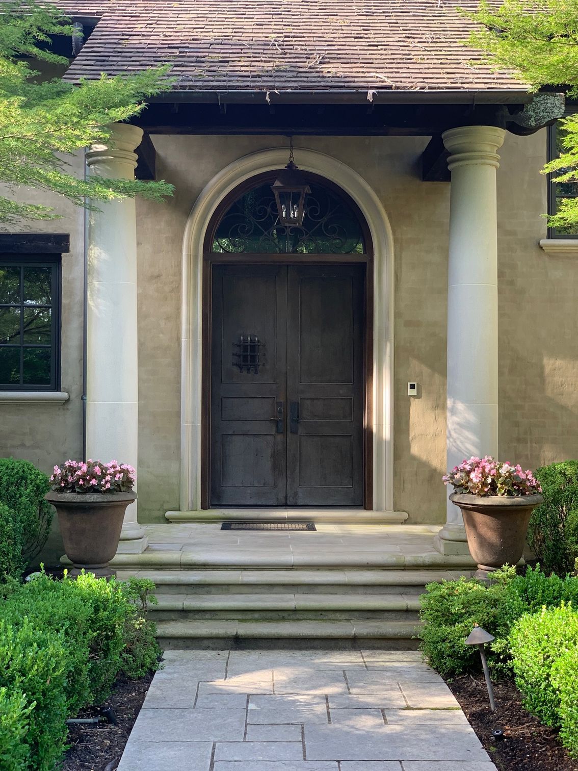 Stone walkway leading to a house with a dark arched doorway flanked by columns, planters, and steps.