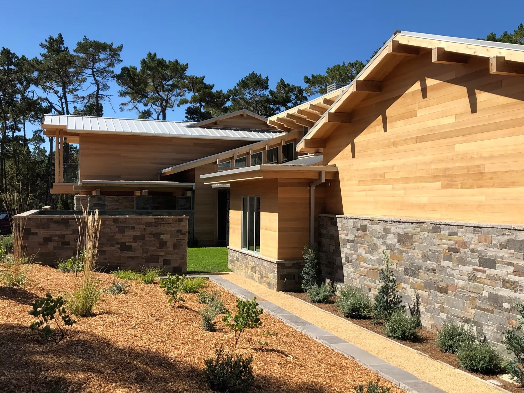 Modern house with wooden siding and stone base, surrounded by landscaping, under a blue sky.