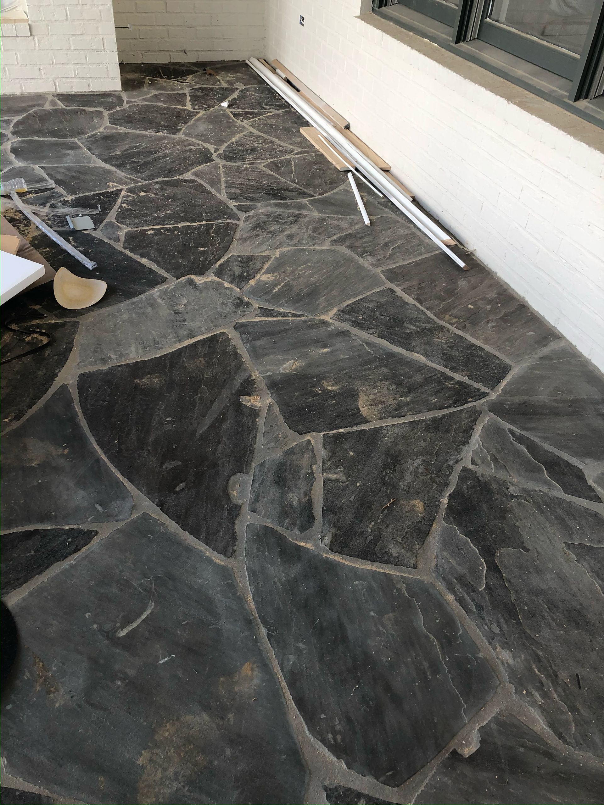 Dark stone patio with irregular shapes and light gray grout, near a white wall and window.