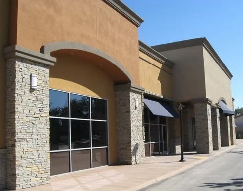 Strip mall storefronts, beige walls, stone columns, large windows, blue awnings, sunny day.