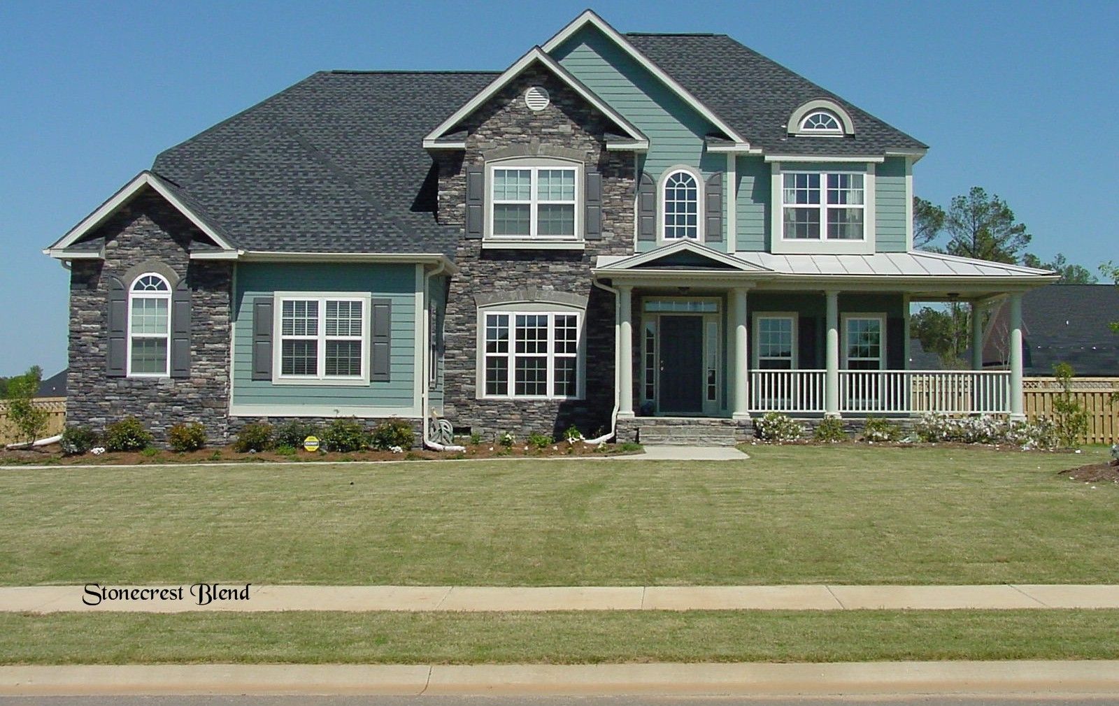 Two-story house with stone and blue siding, front porch, and black roof. Green lawn in front, clear blue sky.