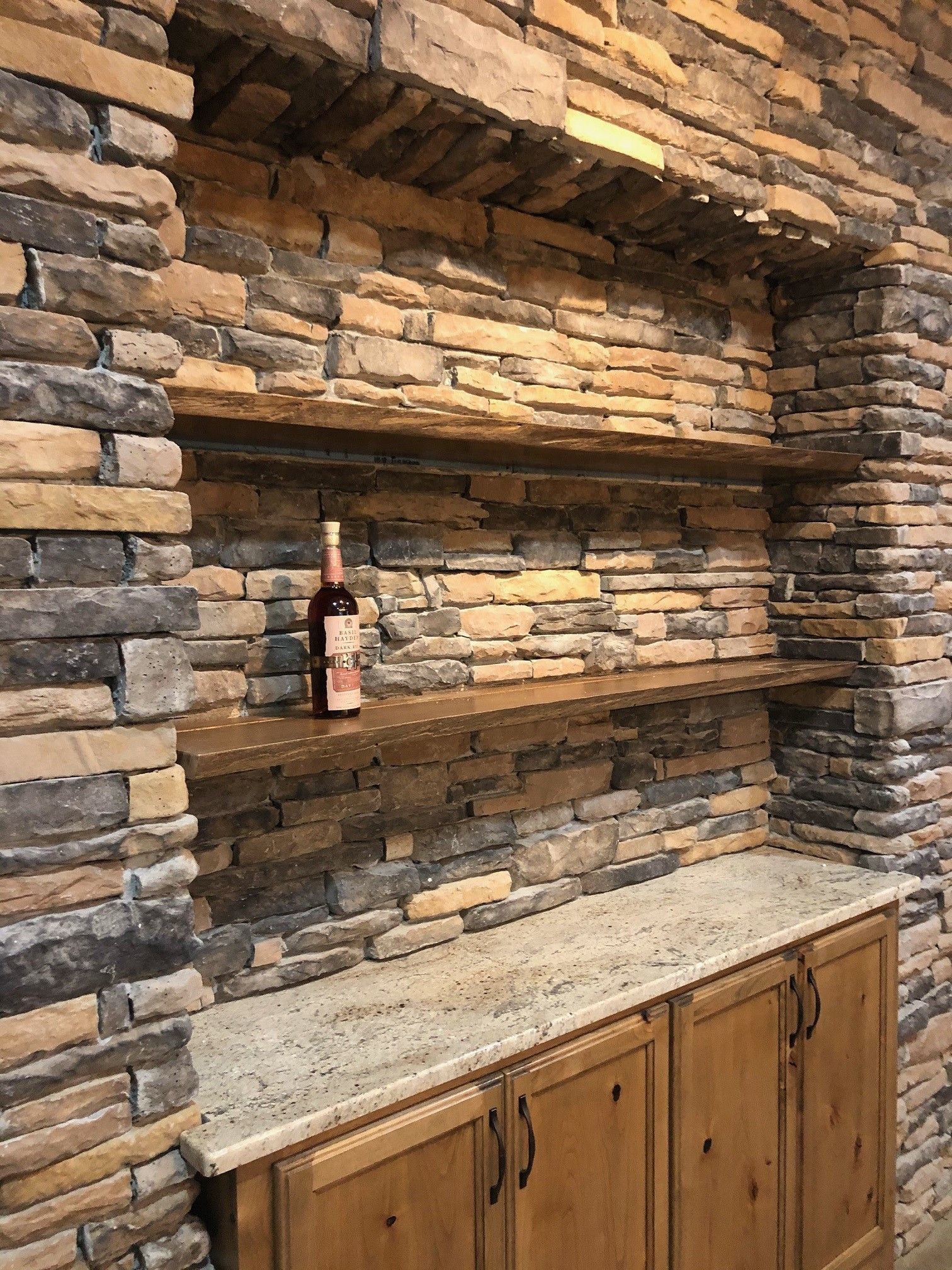 Stone-walled alcove with wood shelves, cabinets, and a granite countertop; wine bottle on middle shelf.