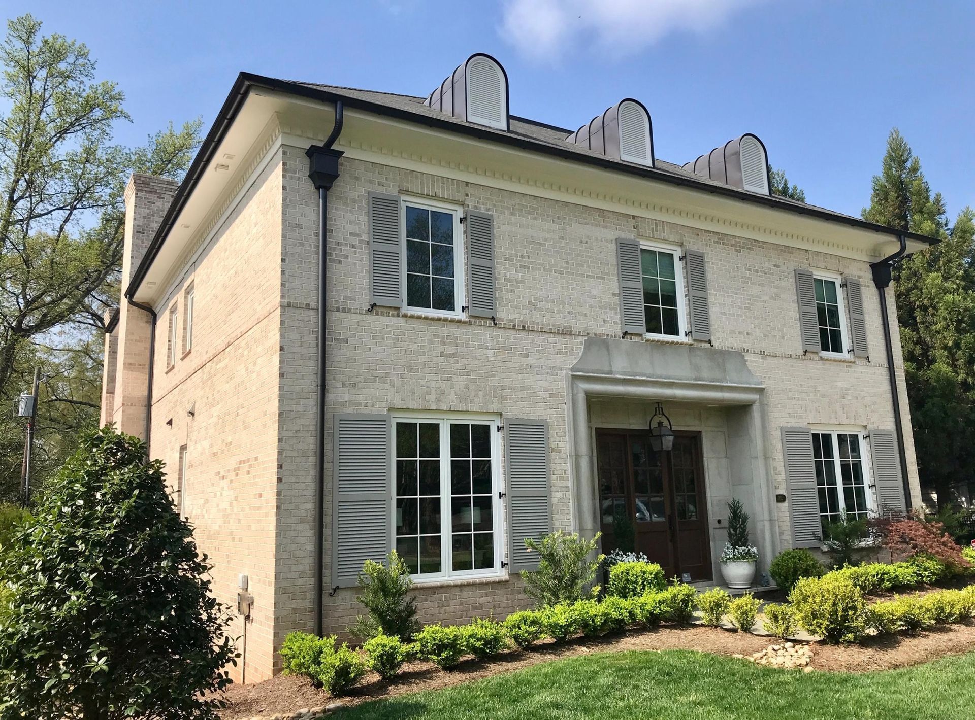 Two-story beige brick house with black roof and gray shutters, front door under small portico, blue sky.