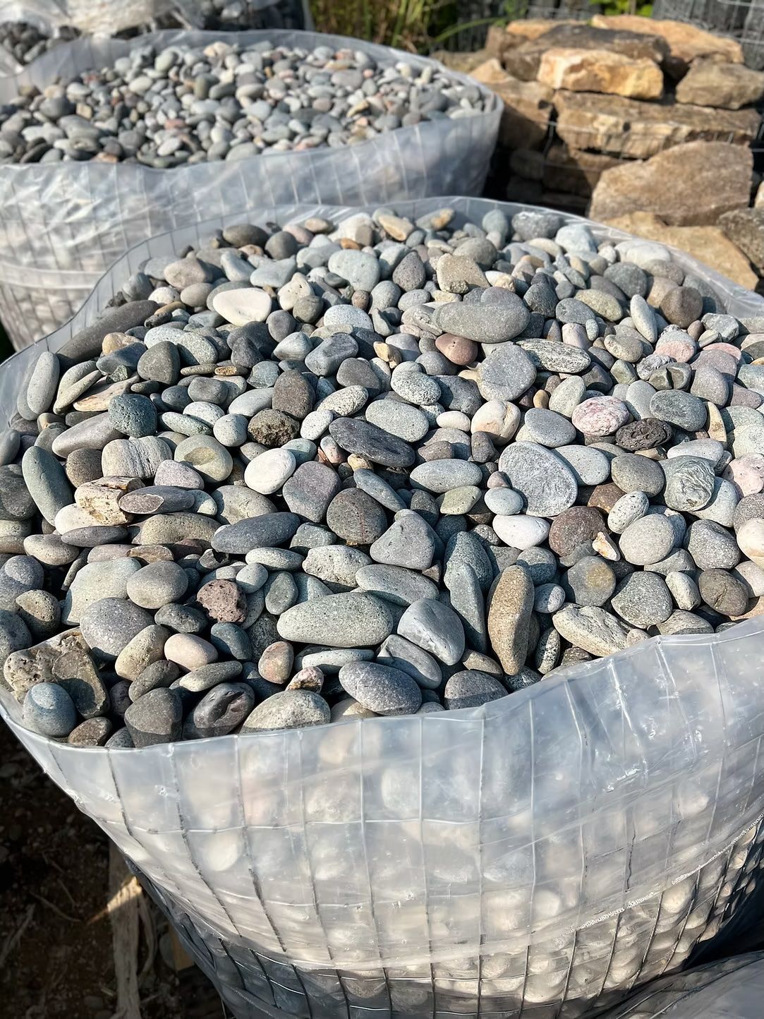 Bags of assorted gray and tan river rocks, likely for landscaping, outside.
