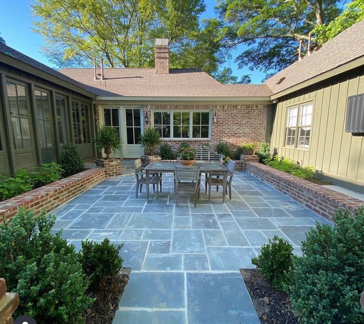 Blue-gray stone patio with a table and chairs, enclosed by brick walls and buildings, plants surround.