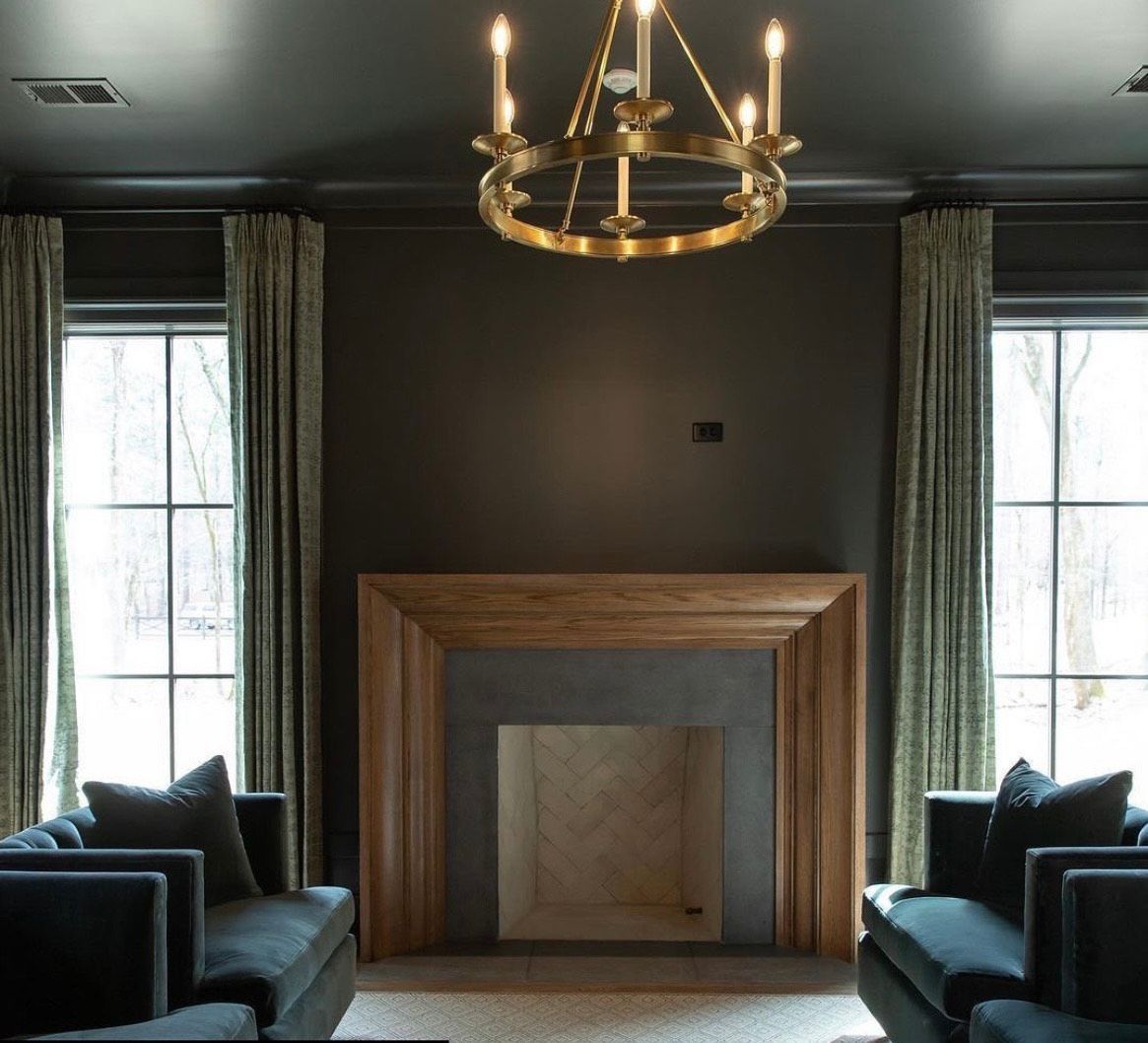 Dark-walled living room with a fireplace, chandelier, windows, and two blue armchairs.