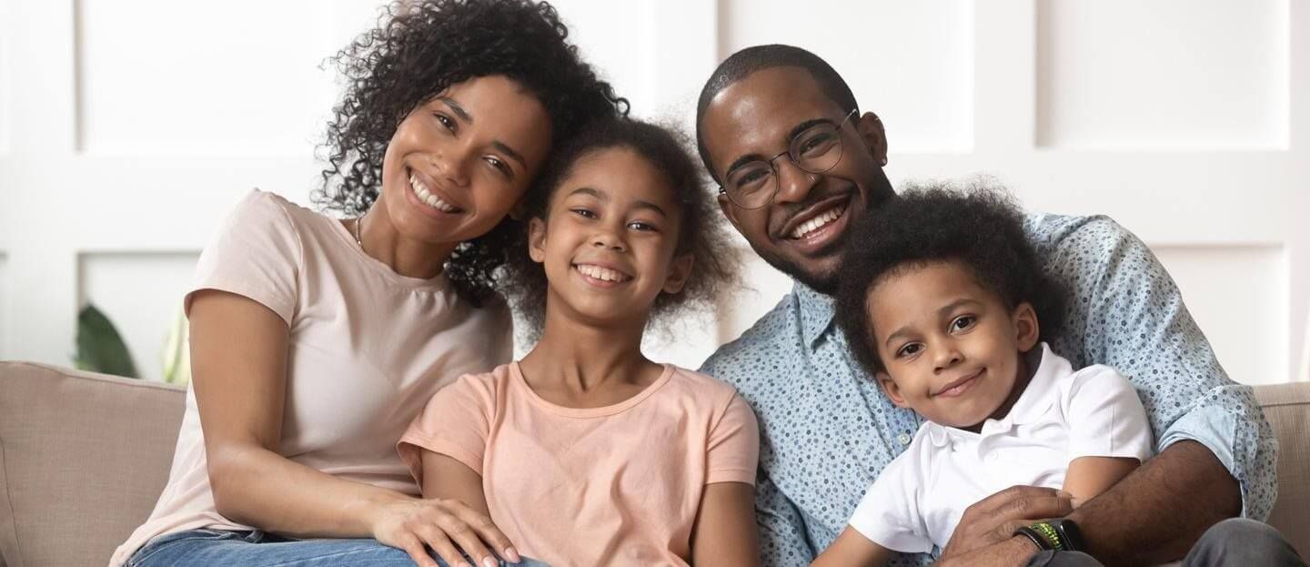 Family of four smiling at the camera, sitting close together on a couch indoors.