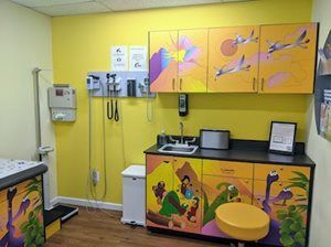 Pediatric exam room with yellow walls, colorful dinosaur artwork, and medical equipment.