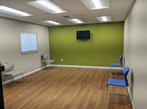 Empty waiting room with blue chairs, TV on green wall, wooden floor, and a window.
