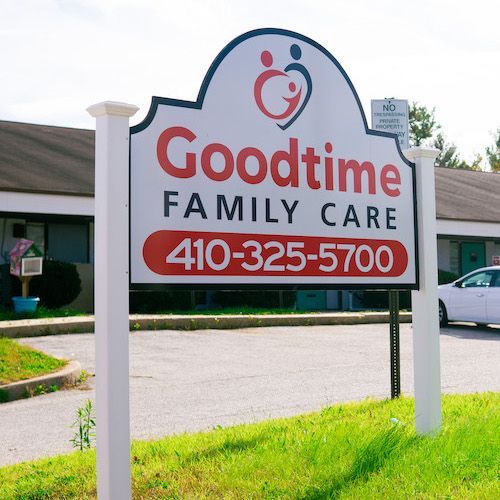 Sign for Goodtime Family Care, red and white logo. 410-325-5700. White posts on green lawn, building in background.