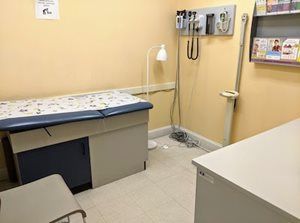 Examination room: examination table, wall-mounted medical equipment, small stool, white cabinet, light beige walls.
