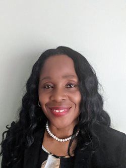 Woman with long black hair, smiling, wearing a pearl necklace and blazer, against a light wall.