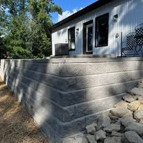Concrete retaining wall beside a house with windows, sloping down to a rocky yard.