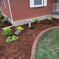 Curved mulch garden bed with small shrubs and a birdbath beside a red house and lawn.