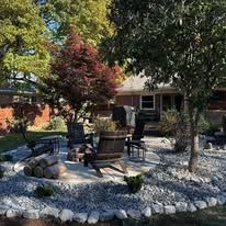 Backyard patio with chairs, stone fire pit, and white rock landscaping under trees