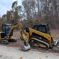 Two yellow mini excavators digging at a roadside in a wooded area.
