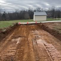 Dirt excavation site with tire tracks, orange cones, and a small white shed in the background under an overcast sky