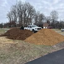Two large dirt piles beside a white pickup truck on a grassy lot with bare trees and houses nearby