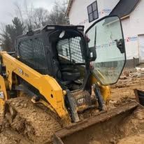 Yellow skid steer stuck in mud on a construction site, with a house in the background