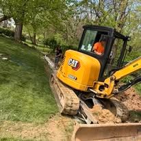 Yellow CAT excavator on a grassy slope near trees, with an operator in the cab.