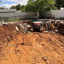 Excavator digging a large dirt lot near a fence and trees under a cloudy sky