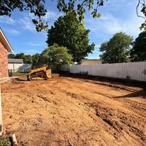 Freshly graded dirt yard beside a brick house, with a small excavator and white fence under blue sky