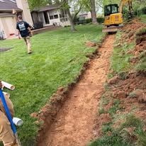 Excavator digging a narrow trench along a grassy yard beside a house