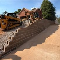 Construction equipment on stepped retaining wall beside a house and dirt driveway