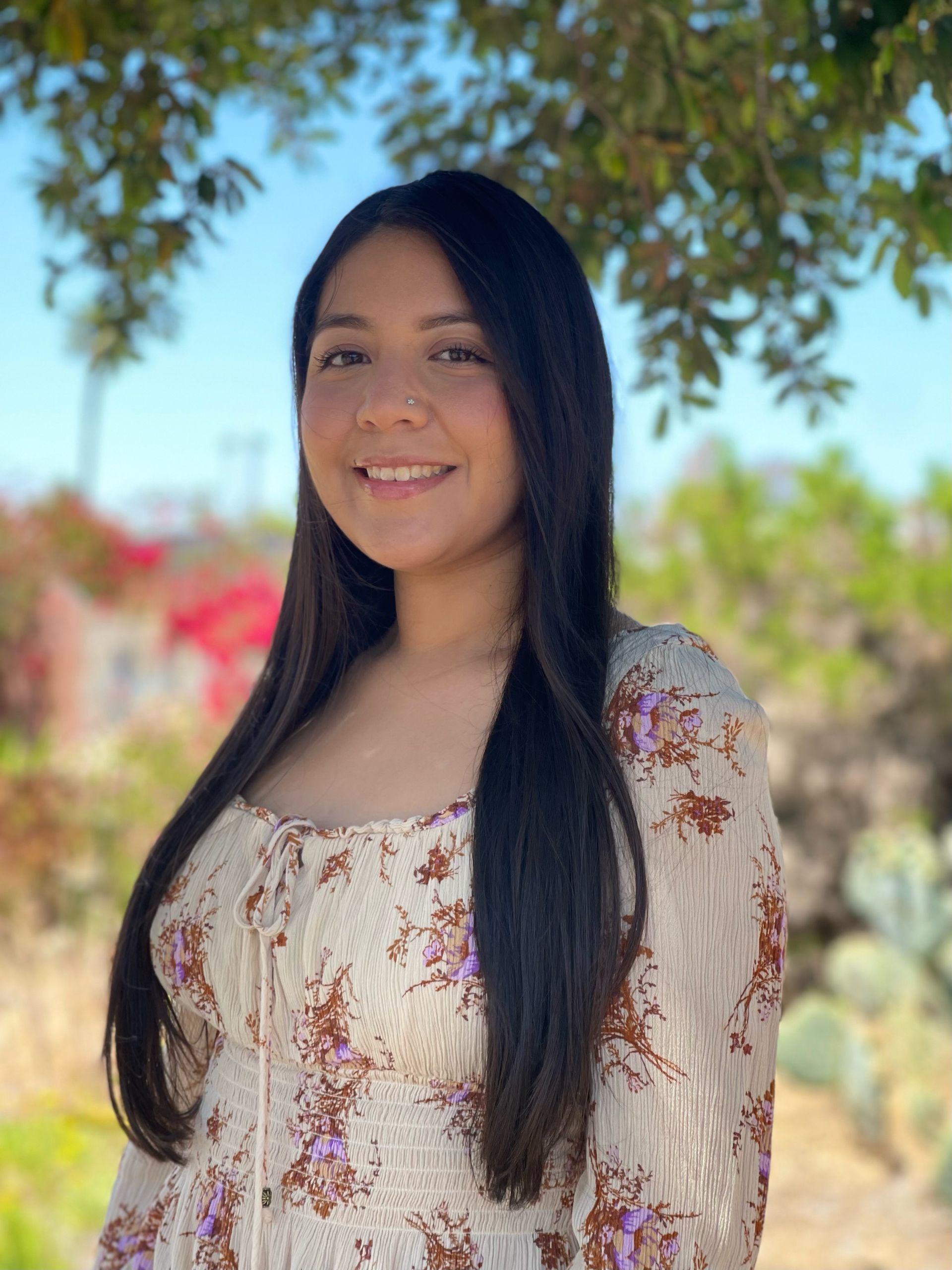A woman with long hair is smiling in front of a bush of pink flowers