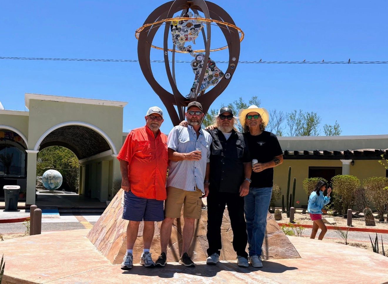 Four people posing outdoors in front of a large metal sculpture and arched building under a blue sky
